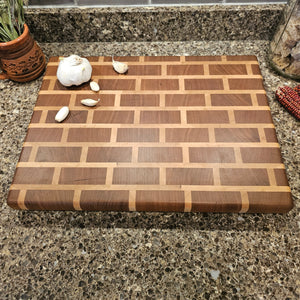 A wooden cutting board with a brick pattern design is placed on a speckled granite countertop. There is a whole garlic bulb and several garlic cloves scattered on the cutting board. To the left, there is a decorative clay pot with dried herbs, and to the right, there is a glass jar with a cork lid and some red decorative items.